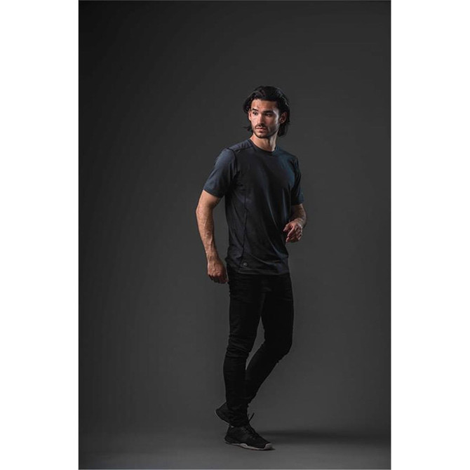 A model wearing a short-sleeve men's T-shirt in dark grey, standing against a black background. The shirt features a logo.