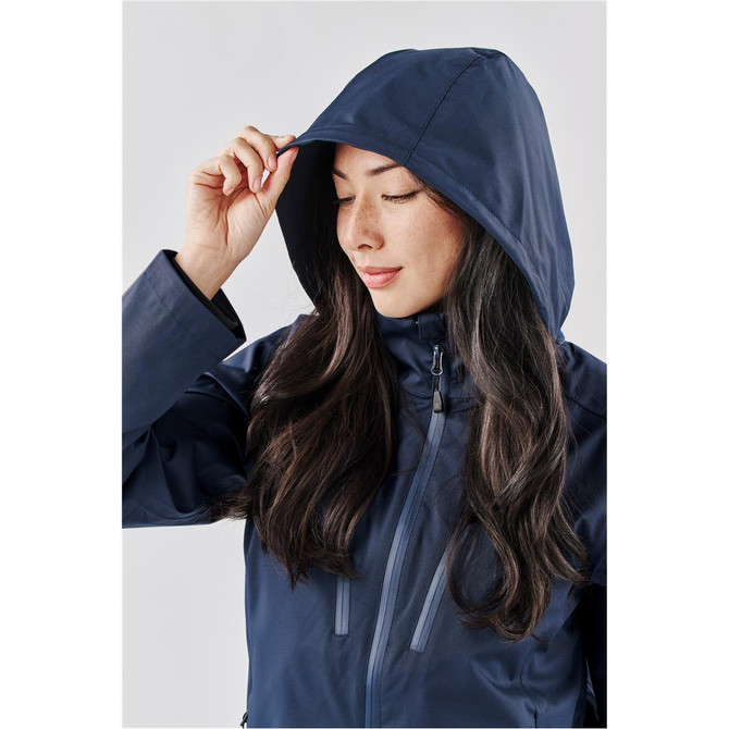 A woman in a navy rain jacket with a hood, adjusting it with one hand. The jacket has a sleek design and a branding logo.