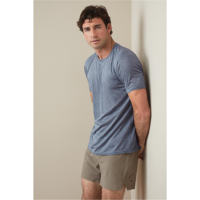A man is wearing a blue recycled sports T-shirt, standing against a light wall, paired with beige shorts.