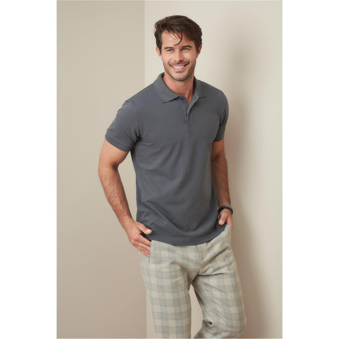 A man wearing a grey polo shirt stands confidently, paired with light-coloured checkered pants, in a neutral setting.