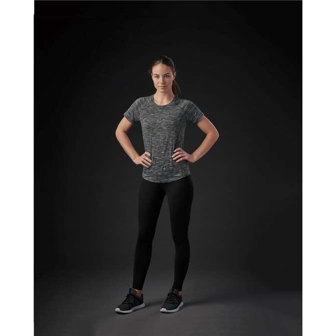 A woman poses in a fitted, multicoloured grey Pacifica tee, paired with black leggings and shoes against a dark background.