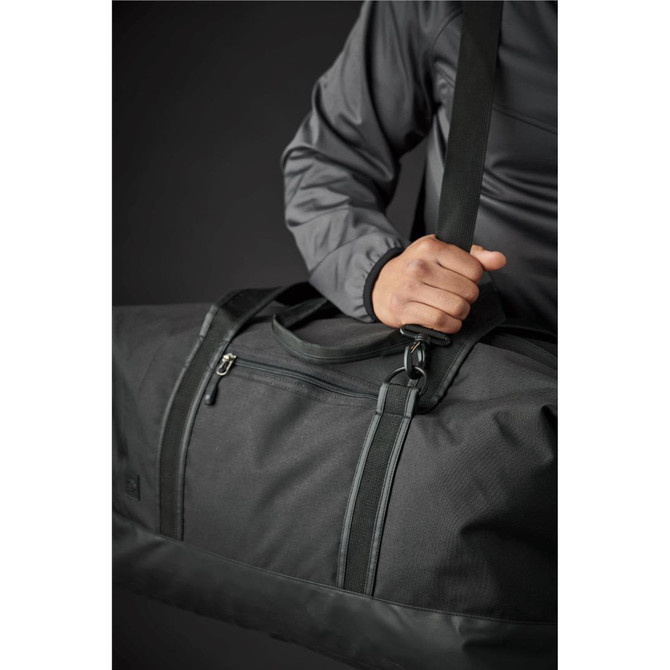 A black duffle bag is being held by a person, showcasing its sturdy handle and sleek design. The bag has a logo.