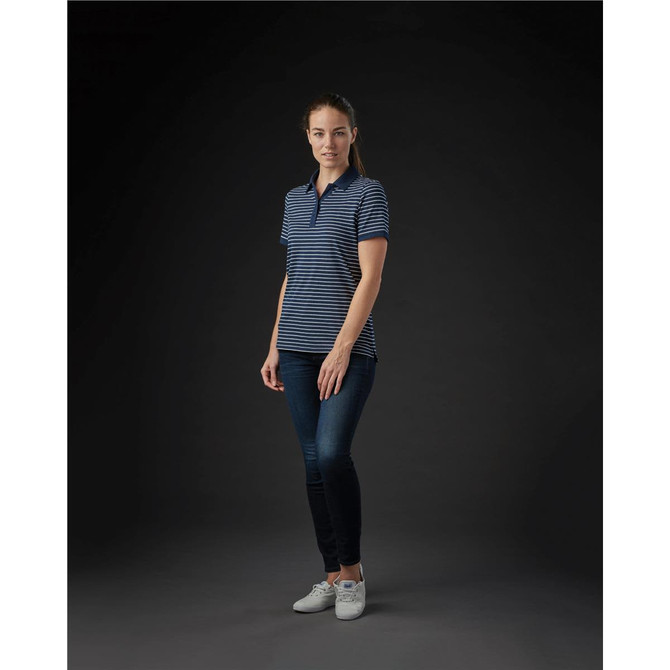 A woman wearing a navy and white striped polo shirt, paired with blue jeans and white shoes, standing against a dark background.