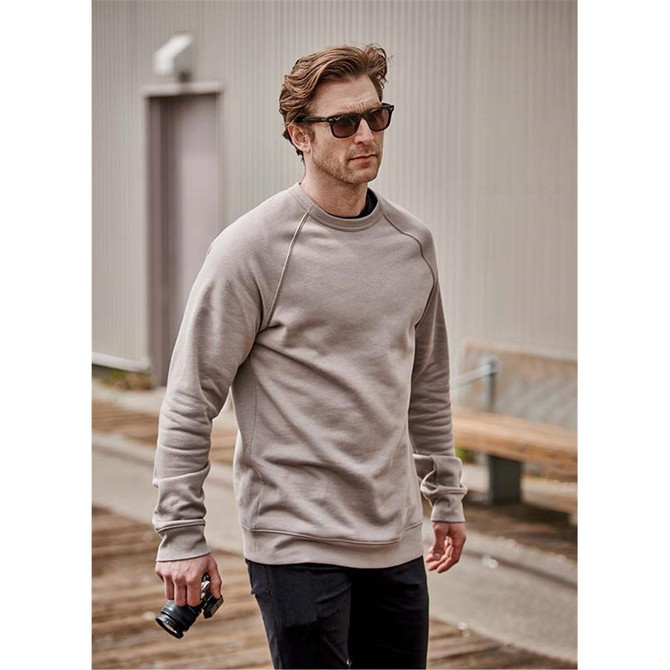 A man wearing a beige crew neck fleece sweater, holding a camera, outdoors in a casual setting.