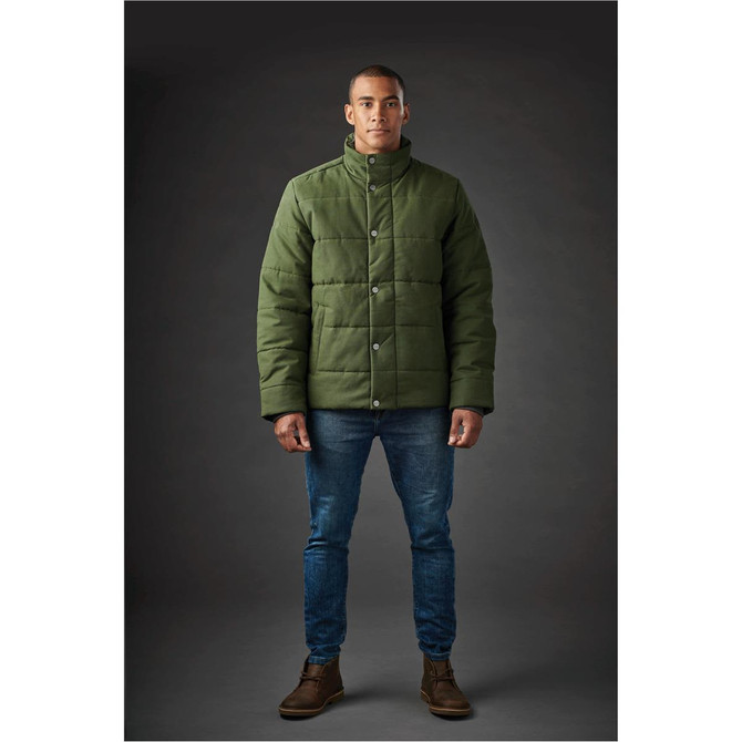 A man stands wearing a dark green thermal jacket with a button-up front, paired with jeans and boots.