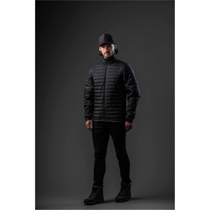 A man wearing a black puffer jacket stands against a dark background, complemented by black trousers and boots.