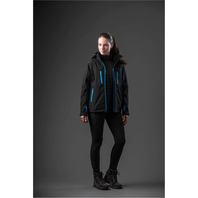 A woman in a black jacket with blue accents stands against a dark background, wearing black leggings and boots.