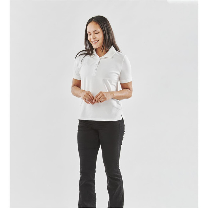 A woman stands wearing a white polo shirt and black pants. The shirt has a logo on it.