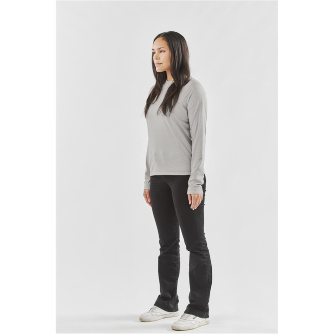 A woman stands in profile wearing a grey long-sleeve tee and black jeans, with white sneakers. The tee features branding.