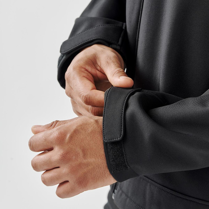 A close-up of a man's hands adjusting the cuffs of a black softshell jacket with velcro closures.