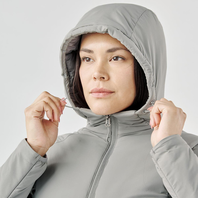A woman wearing a light grey thermal puffer jacket with a hood, adjusting it around her face.