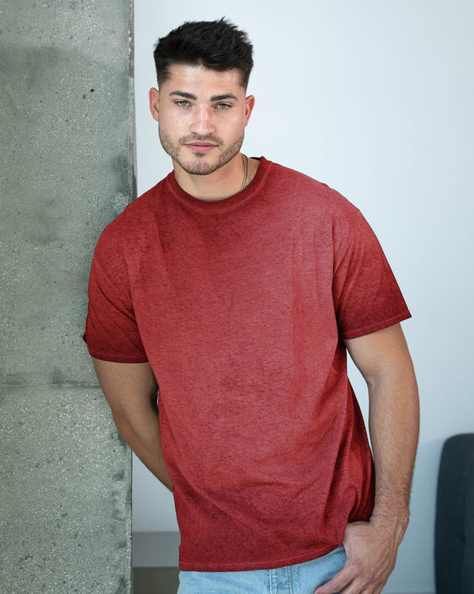 A man leans against a concrete pillar, wearing a maroon Colortone® Oil Wash T-Shirt. The shirt has a casual, relaxed fit.
