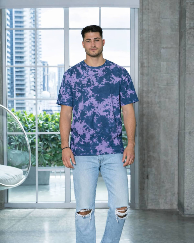 A man stands in a modern space wearing a purple and navy tie-dye T-shirt and ripped jeans. The shirt features a logo.