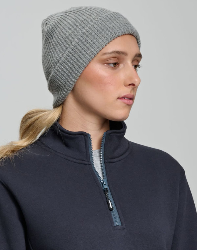 A grey cable knit beanie displayed on a model wearing a navy zip-up top, featuring a logo on the beanie.