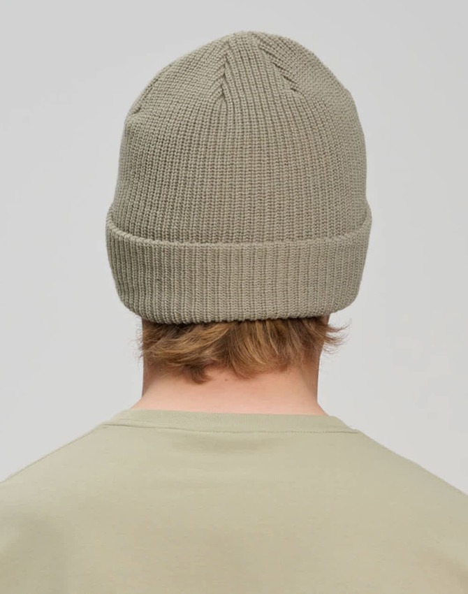 A roll-up cable knit snug beanie in a light khaki colour, viewed from the back. Features a ribbed band and a logo.