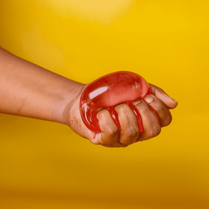 A hand squeezing a translucent red jelly cube sensory toy against a bright yellow background.