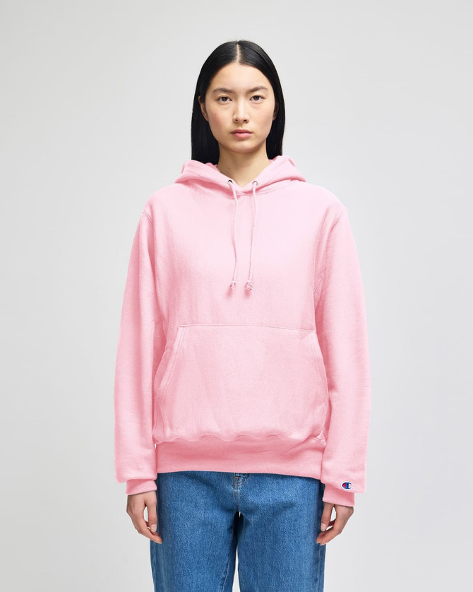 A pink pullover hooded sweatshirt with a front pocket and a branded logo. Model is wearing it with blue jeans.