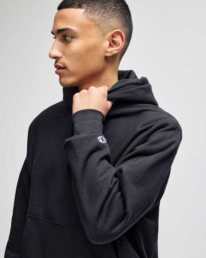 A black pullover hooded sweatshirt with a logo, worn by a model, showing the hood and sleeve.