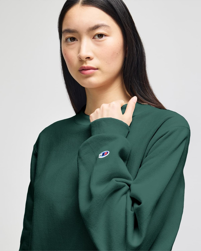 A model wearing a dark green Champion Reverse Weave® adult crew sweatshirt featuring a logo on the sleeve.