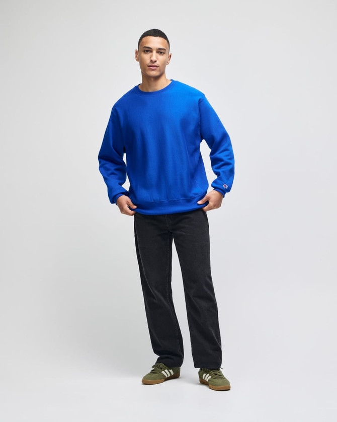 A blue Champion Reverse Weave® adult crew sweatshirt worn with black pants and green shoes, featuring a logo on the sleeve.