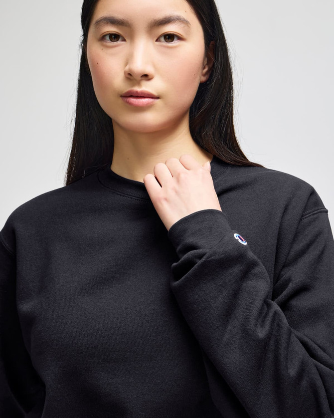 A black crewneck sweatshirt featuring a logo on the sleeve, worn by a person with long hair.