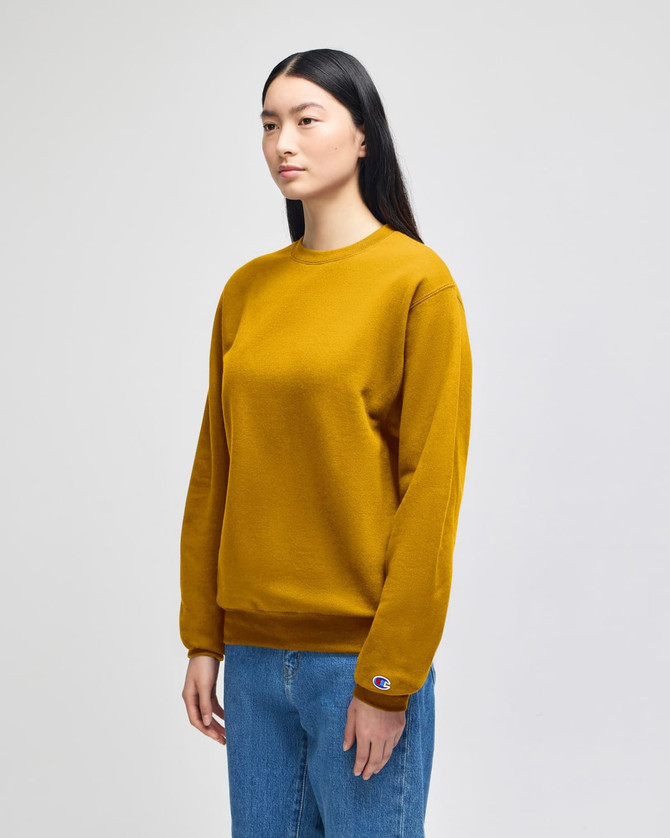 A mustard yellow crewneck sweatshirt is displayed on a model, featuring long sleeves and a logo on the cuff.