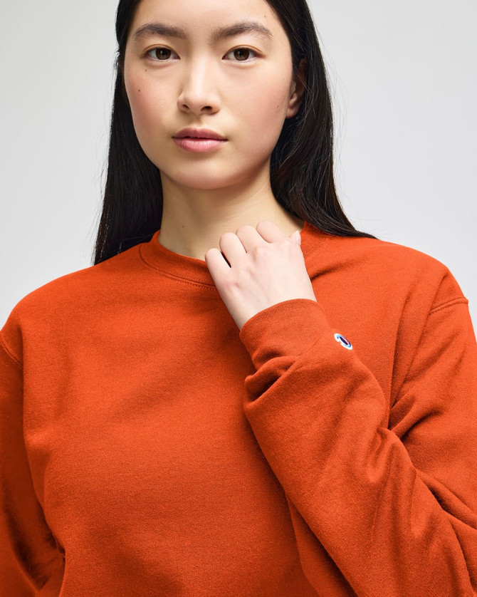 An adult crewneck sweatshirt in orange worn by a model, featuring a logo on the sleeve.