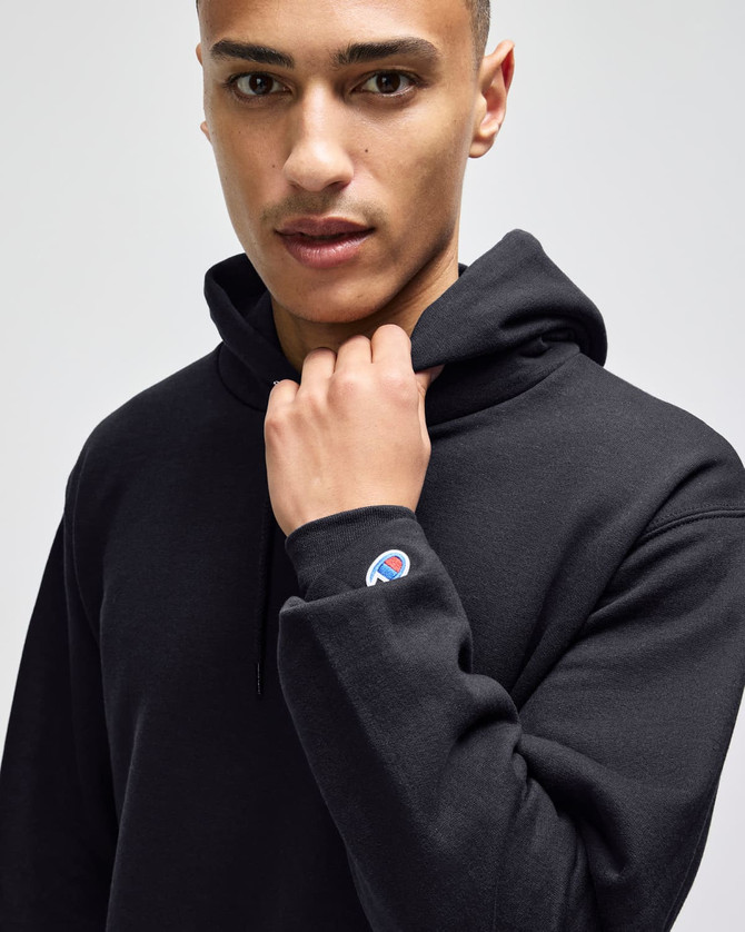 A black pullover hooded sweatshirt is worn by a model, featuring a small logo on the sleeve.