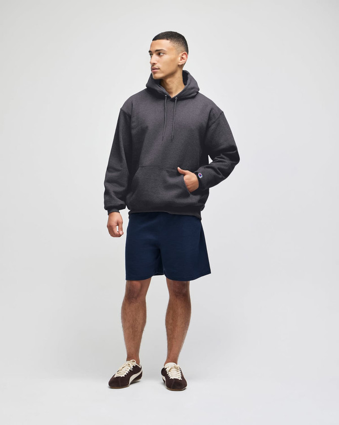 A model wears a grey hooded sweatshirt paired with navy shorts and shoes. The sweatshirt has a front pocket and a logo.