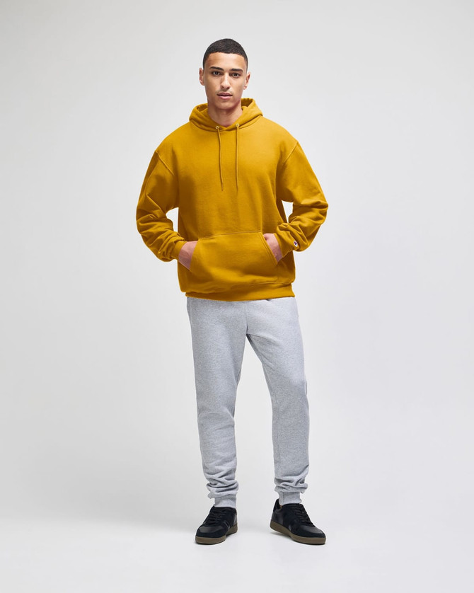 A mustard yellow pullover hooded sweatshirt worn by a model with grey track pants and black shoes. The sweatshirt has a logo.