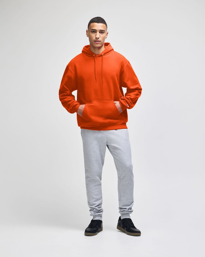 An orange pullover hooded sweatshirt is worn with grey track pants, complemented by black shoes.