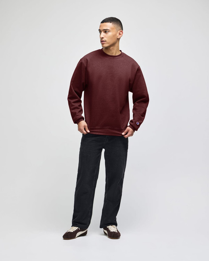 A man stands in a burgundy pullover hooded sweatshirt and dark pants, featuring a logo on the sleeve.