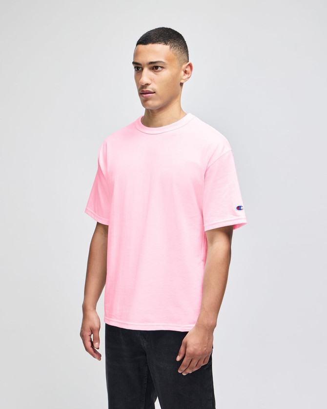 A pink Champion Adult Heritage Jersey T-shirt displayed on a model, featuring a logo on the sleeve.