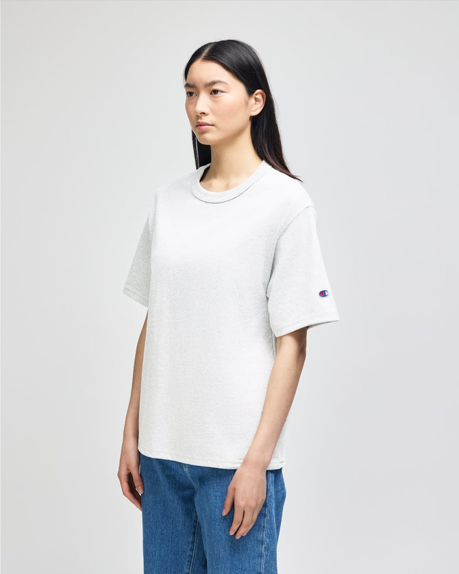A white Champion adult heritage jersey t-shirt on a model, featuring a logo on one sleeve.