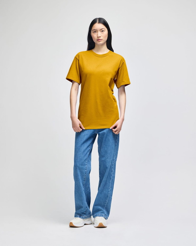 A model wearing a mustard yellow Champion adult short-sleeve T-shirt paired with blue jeans. The shirt features a logo.