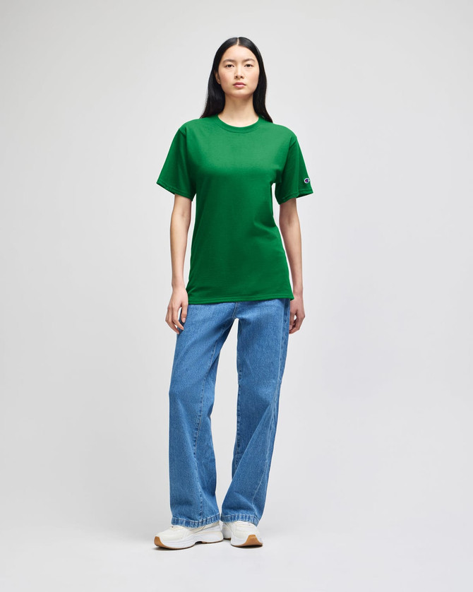 A green short-sleeve t-shirt is displayed on a model paired with blue jeans and white sneakers. It features a logo on the sleeve.