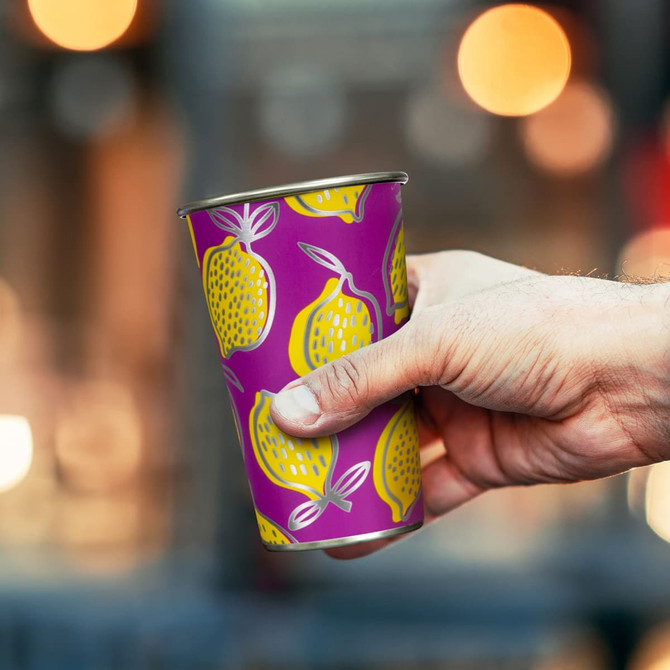 A drink cup featuring a vibrant purple design with yellow lemons and a silver rim, held in a hand.