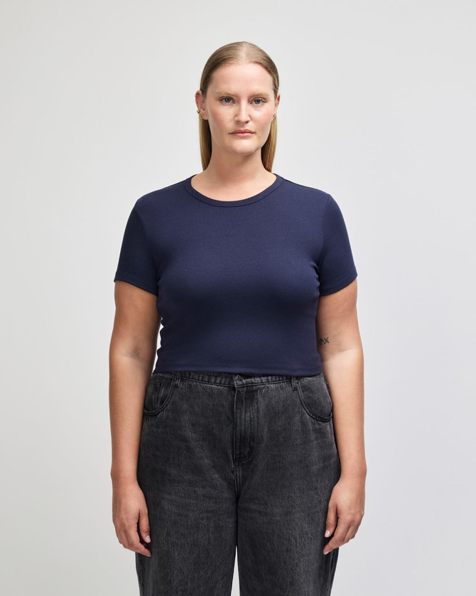 A navy blue crop fitted t-shirt on a model, paired with black high-waisted jeans. The shirt has a logo.