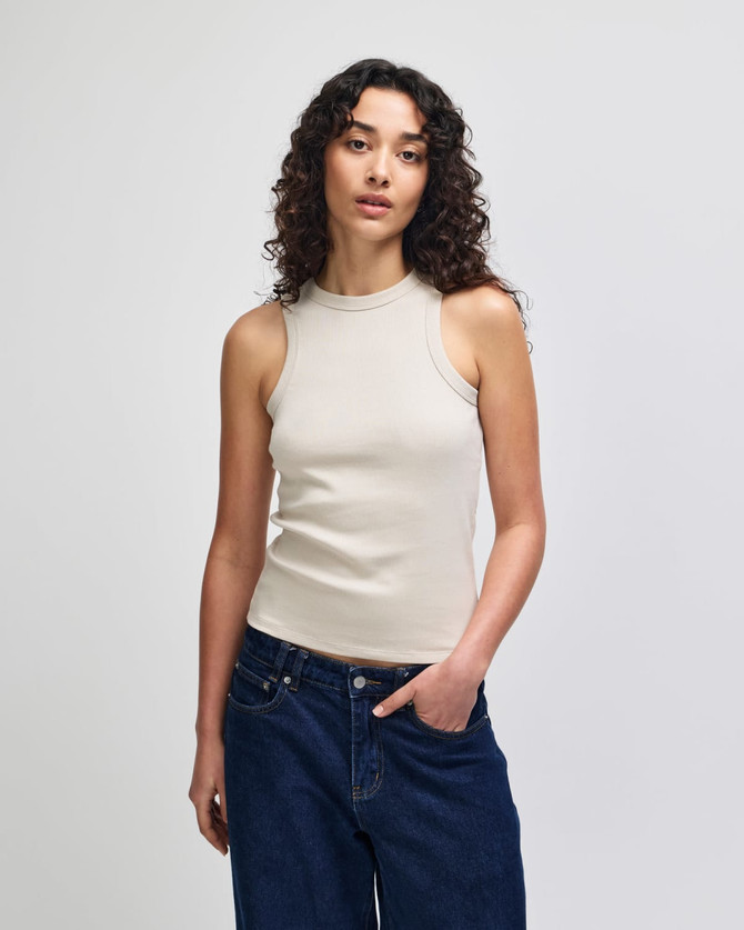 A fitted tank top in a neutral beige colour, worn by a model with curly hair, paired with blue jeans.