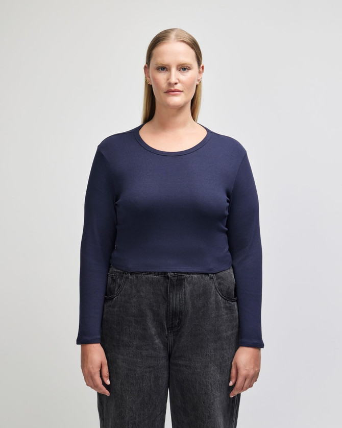 A long-sleeved, fitted crop top in navy blue, paired with high-waisted dark jeans. The top features a round neckline and has a logo.