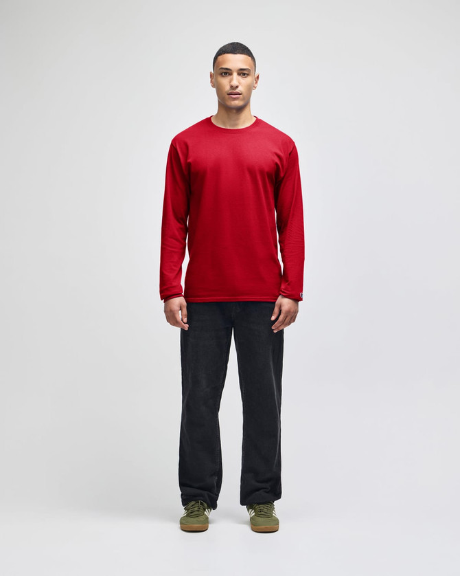A red long-sleeve t-shirt on a model, paired with dark trousers and green shoes. The shirt features a logo.
