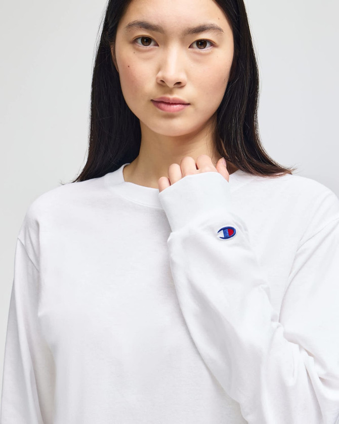 A white long-sleeve t-shirt featuring a logo on the sleeve, modelled by a person with long dark hair.