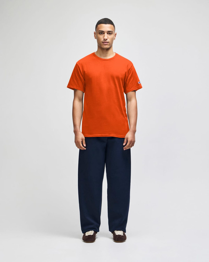 An orange short-sleeve t-shirt worn by a man, paired with navy blue pants, displaying a logo on the shirt.