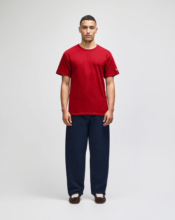 A red short sleeve t-shirt on a model, paired with navy pants, featuring a subtle logo on the sleeve.
