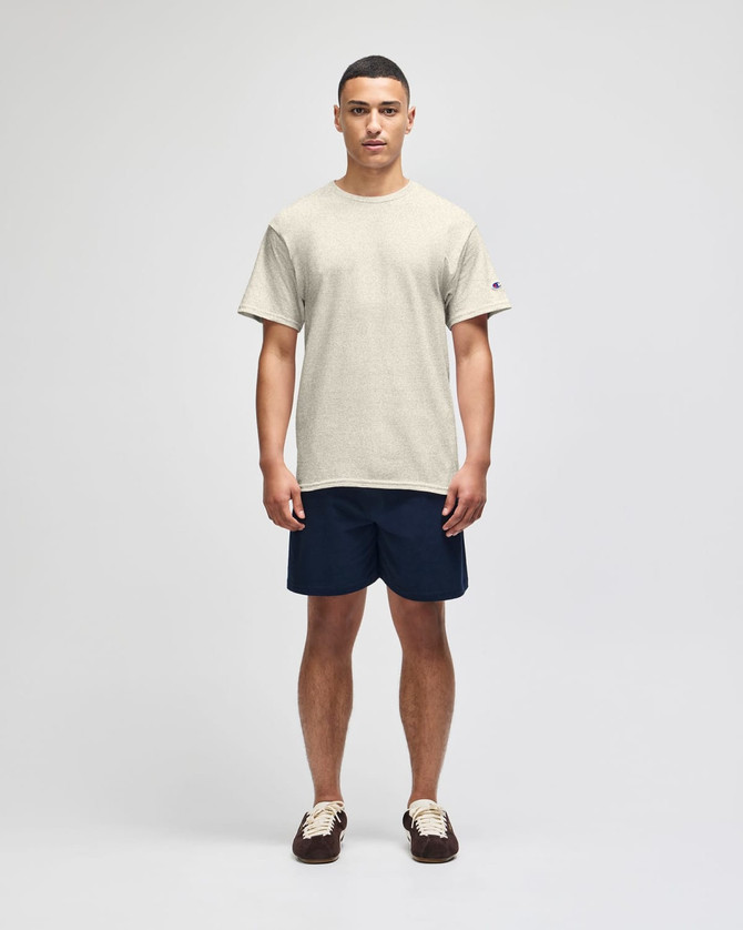 A beige short sleeve t-shirt displayed on a male model, paired with navy shorts. The shirt features a logo on the sleeve.