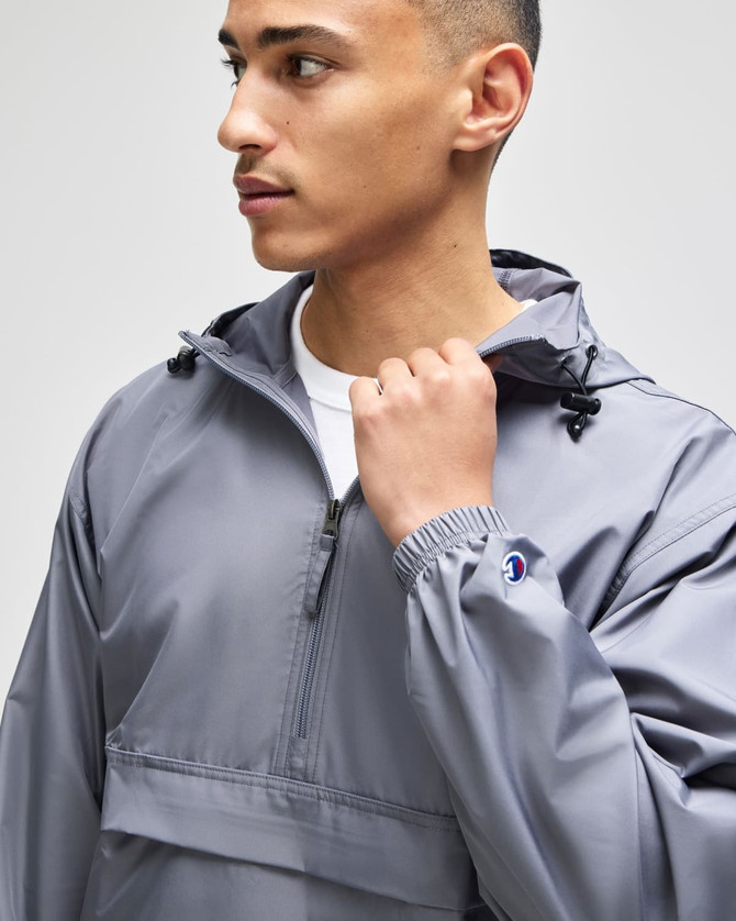A grey packable anorak jacket with a hood, featuring a front pocket and logo on the cuff.