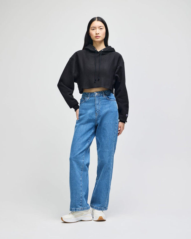A woman wearing a black cropped hoodie with loose blue jeans and white sneakers, standing against a plain background.