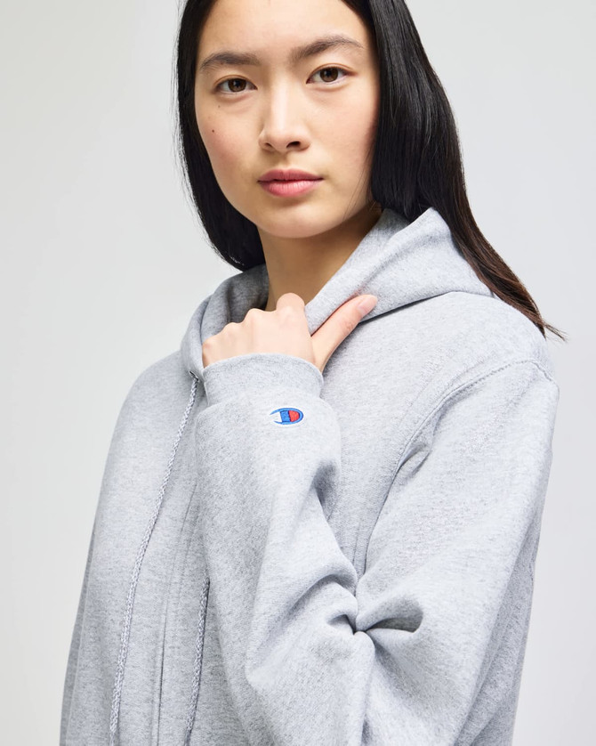 A woman wearing a grey Champion Powerblend® adult full-zip hooded sweatshirt, featuring a logo on the sleeve.
