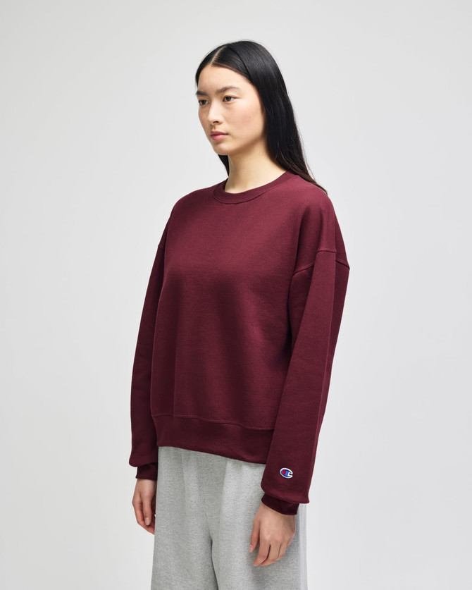 A maroon women's crewneck sweatshirt with a relaxed fit, featuring a logo on the sleeve. Styled with grey pants.
