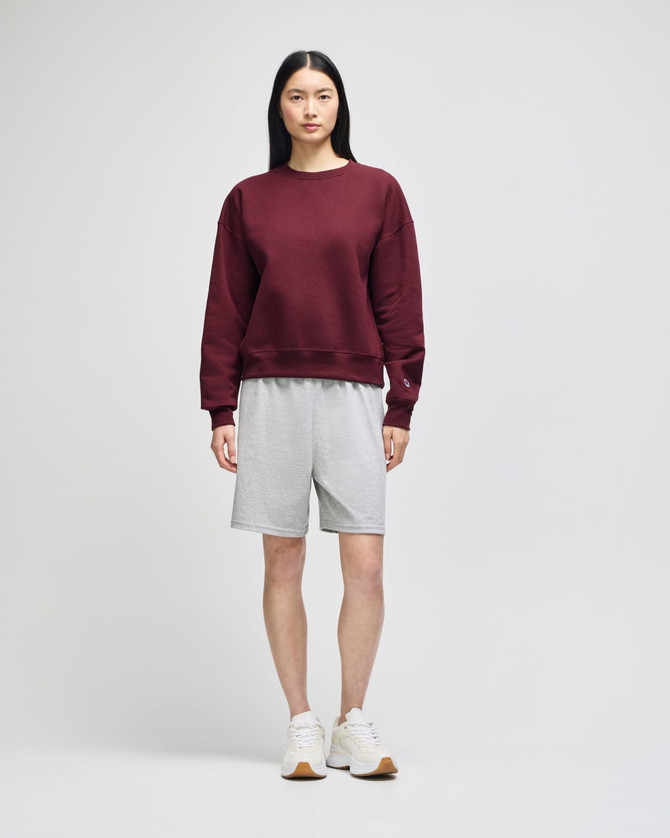 A burgundy crewneck sweatshirt paired with grey shorts on a model, with white sneakers. The sweatshirt features a logo.